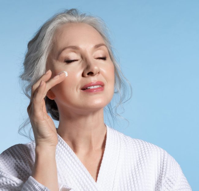 Senior woman with eyes closed putting cosmetic moisturizing rejuvenating cream on face feeling pleasure. Studio portrait on blue. Refreshment and anti-wrinkle skin therapy